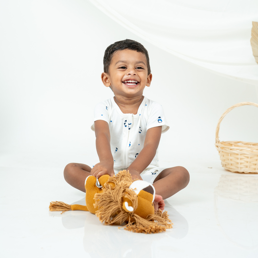 A smiling toddler boy sits cross-legged on a white studio floor, holding a plush lion toy. He is wearing a white short-sleeve bamboo romper with small blue penguin cartoon motifs.