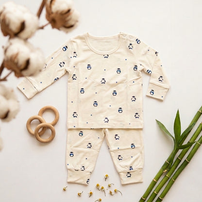 A baby long-sleeve bamboo lounge set laid flat on a white background, featuring a cream-colored top and matching pants with a small blue penguin print. The set is styled with cotton bolls, wooden rings, and green bamboo leaves.