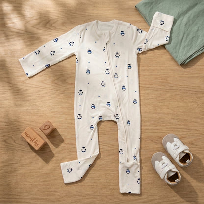 A baby long-sleeve bamboo sleepsuit in off-white with a navy blue penguin pattern, laid flat on a light wood background with baby accessories including wooden blocks, a mint green cloth, and baby shoes.
