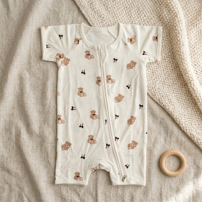A baby short-sleeve footless bamboo romper in off-white, featuring a print of teddy bears and small bows, laid flat on a textured beige fabric next to a wooden teething ring.