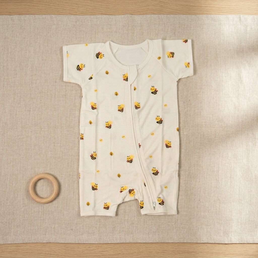 A baby short-sleeve bamboo romper in off-white, printed with a pattern of yellow bumblebees and small yellow flowers, laid flat on a beige linen mat with a wooden ring toy nearby.