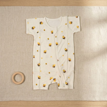 A baby short-sleeve bamboo romper in off-white, printed with a pattern of yellow bumblebees and small yellow flowers, laid flat on a beige linen mat with a wooden ring toy nearby.