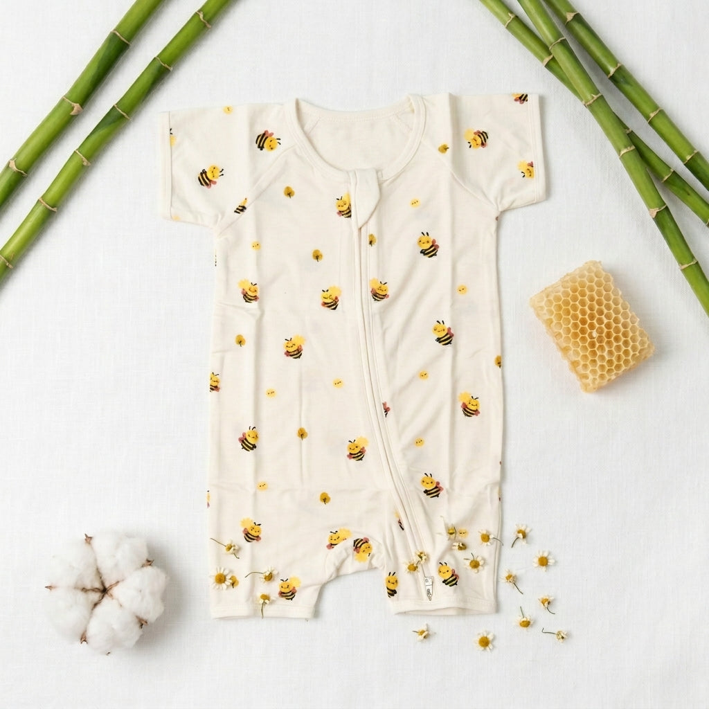 A baby bamboo romper in cream-colored cotton fabric with a repeating pattern of yellow and black bumblebees, styled in a flat-lay with bamboo stalks, a cotton boll, and honeycomb on a white background.