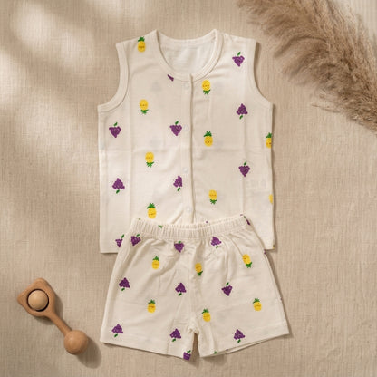 A baby bamboo jabla and shorts set laid flat on a beige textured fabric background. The set is off-white with a repeating pattern of yellow pineapples and purple grapes.