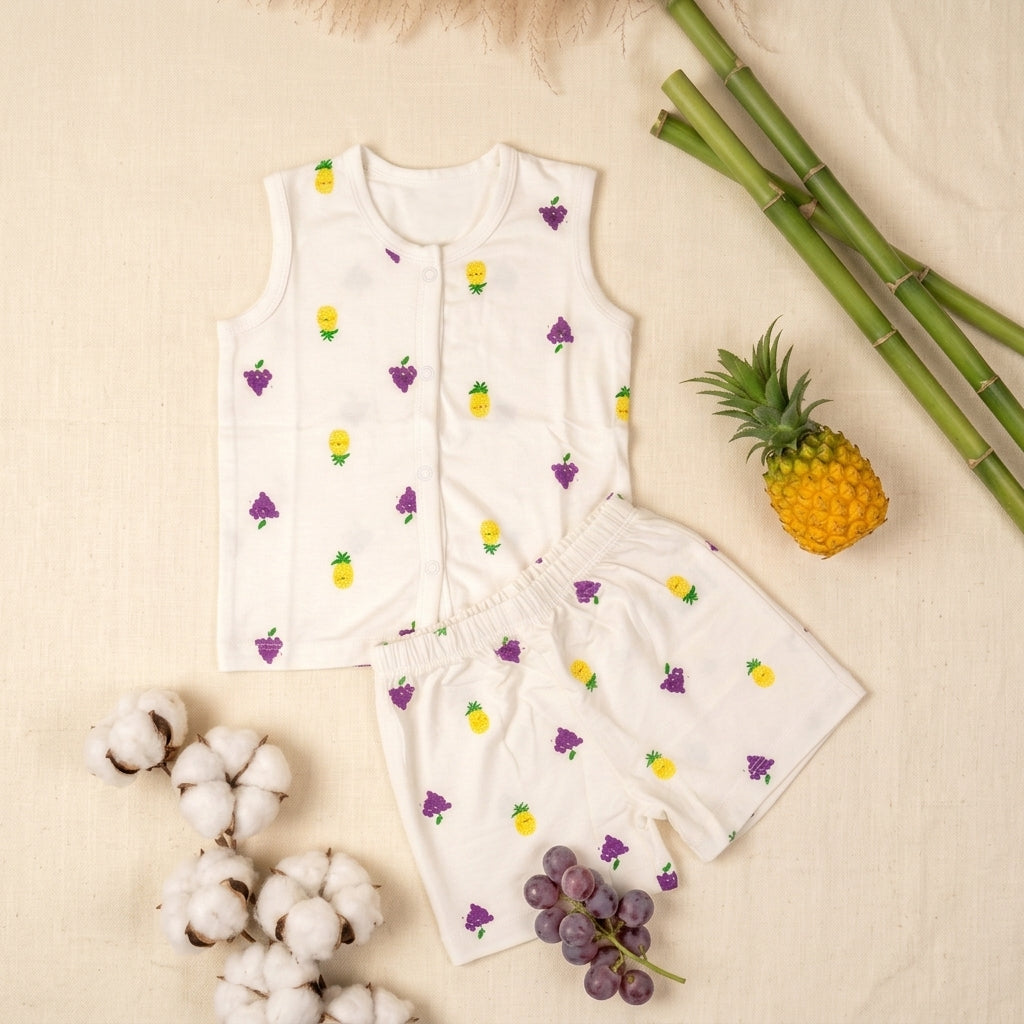 A baby two-piece outfit set consisting of a bamboo jabla and shorts set, decorated with a colorful pattern of yellow pineapples and purple grapes. The set is styled in a flat-lay arrangement on a beige fabric background, surrounded by decorative props including cotton bolls, a pineapple, and a bunch of green bamboo.