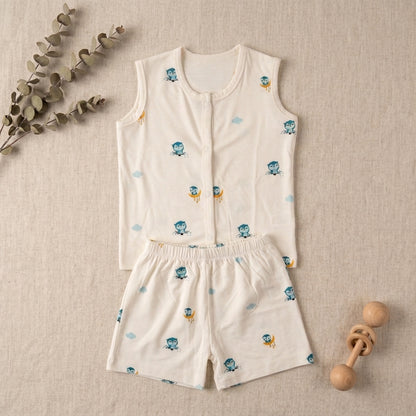 A baby bamboo jabla and shorts in a warm off-white fabric, decorated with a small blue and yellow owl pattern. The set is laid flat on a textured beige background with decorative eucalyptus branches and a wooden baby rattle.