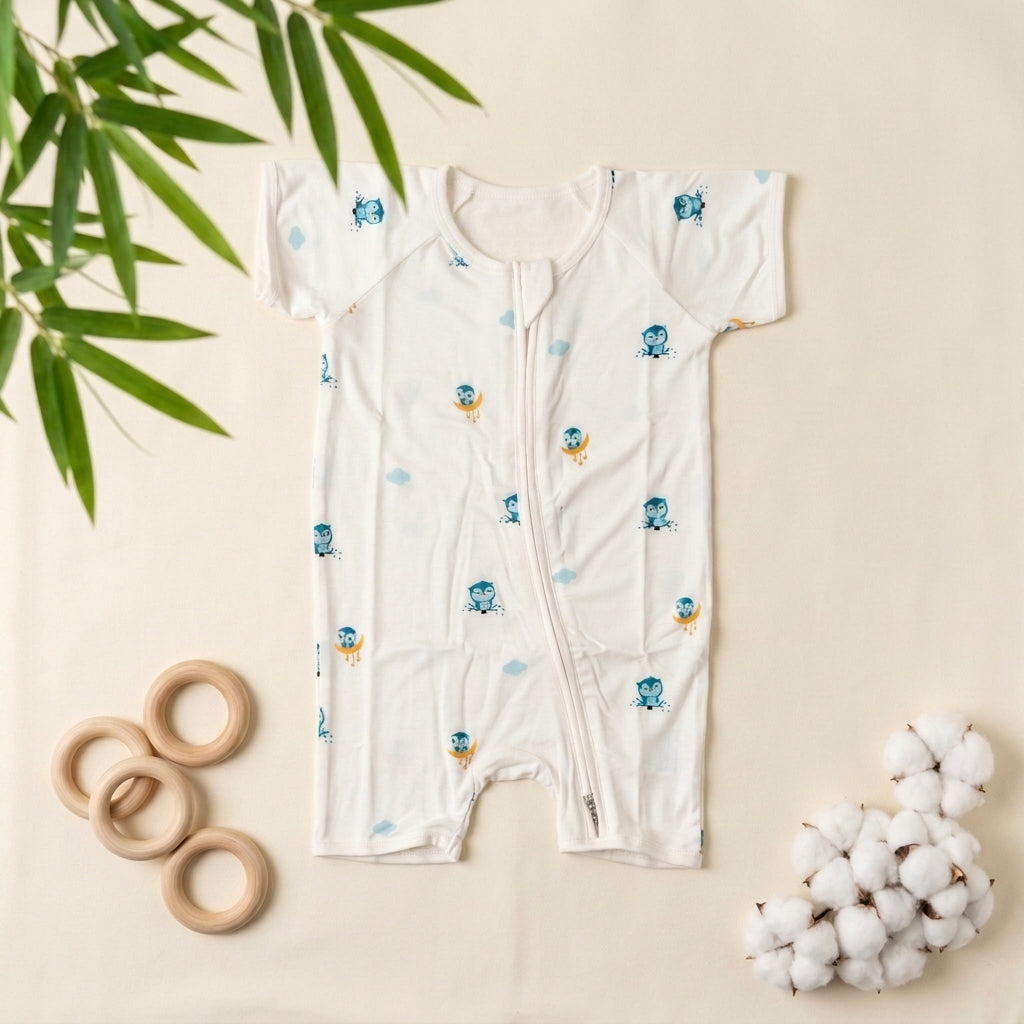 A baby short-sleeve bamboo romper laid flat on a cream background, decorated with a blue octopus and yellow owl pattern. Styled flat-lay props include wooden rings, cotton bolls, and green leaves.