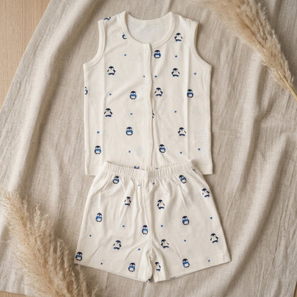 A two-piece baby bamboo jabla and shorts set featuring a sleeveless vest and matching shorts in off-white, decorated with a small blue penguin and star pattern, laid flat on a beige linen fabric with dried pampas grass accents.