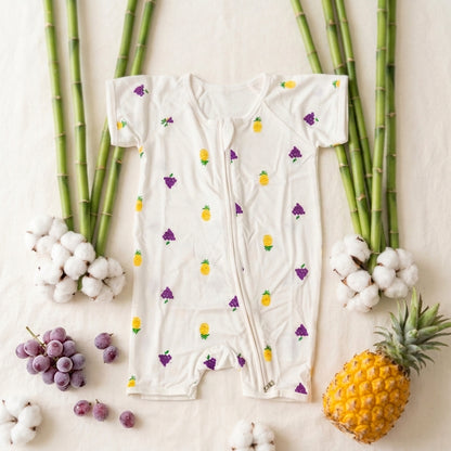 A baby short-sleeve footless bamboo romper in off-white, decorated with a pattern of yellow pineapples and purple grapes. The romper is laid flat on a light beige fabric background, surrounded by decorative props including green bamboo stalks, white cotton bolls, purple grapes, and a whole pineapple.